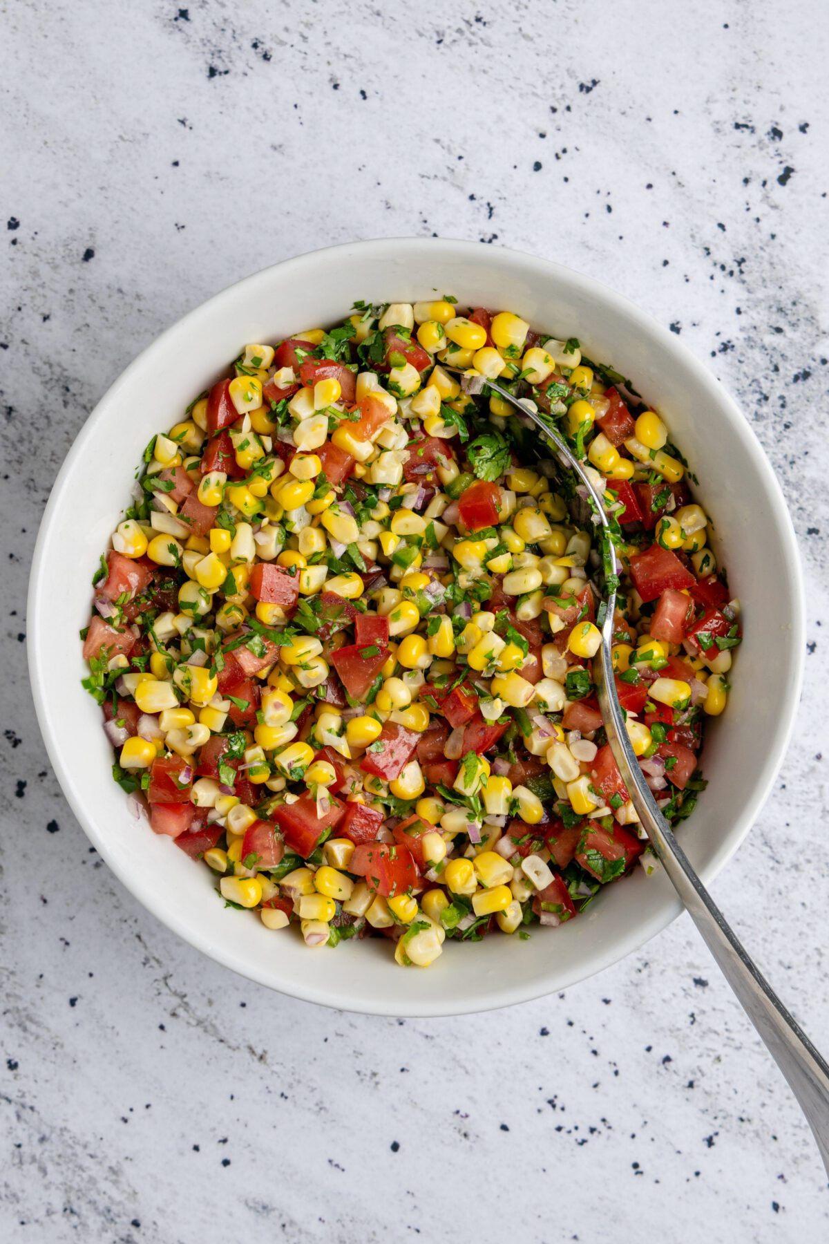 corn, tomatoes, onion, cilantro, jalapeño, lime juice, lemon juice, vegetable oil, and salt combined together in white bowl with spoon