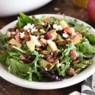 Green salad on white plate with roasted pumpkin seeds, chopped apples, candied pecans, feta cheese and red onions.