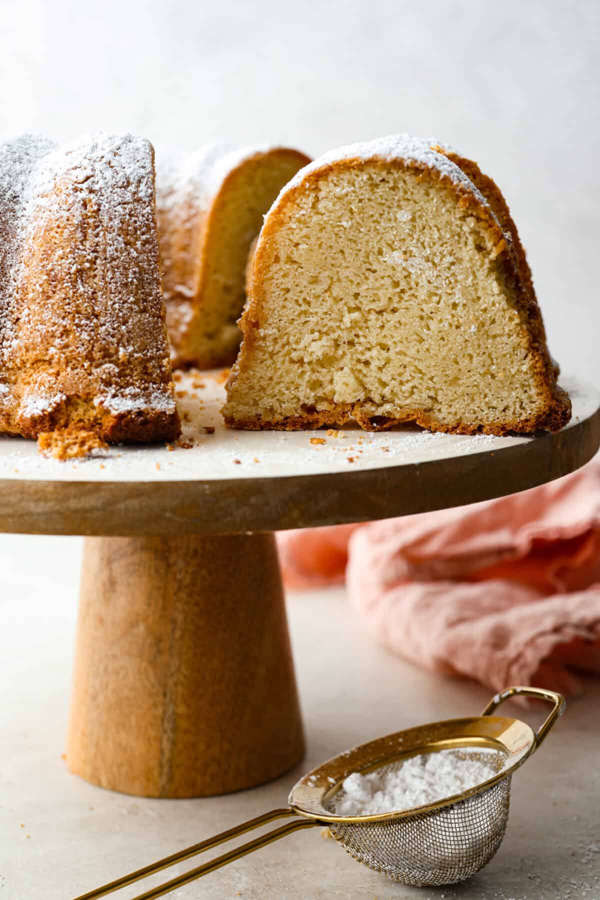 Sour cream pound cake on a cake stand with a tine gold sifter with powdered sugar in it in the foreground. 