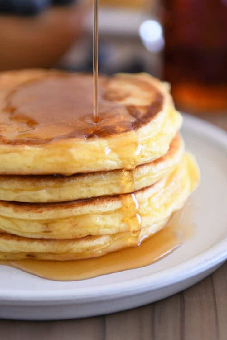 Drizzling pure maple syrup over flour stacked cottage cheese pancakes on gray plate.