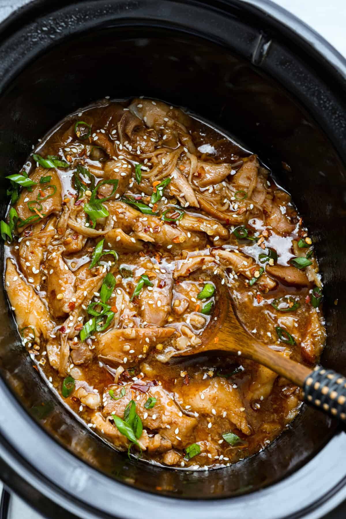 Slow cooker Korean BBQ chicken in a crockpot with a wooden spoon, with sesame seeds and green onions. 
