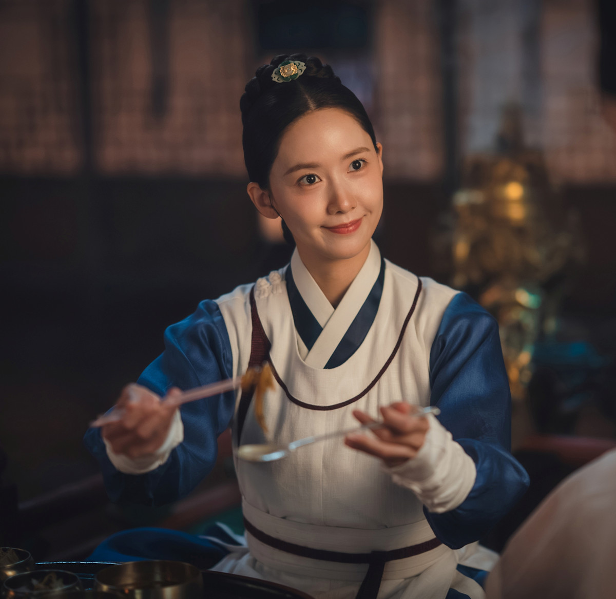 A smiling woman in ancient white and blue Korean garb smiles as she offers up a bite of food with chopsticks