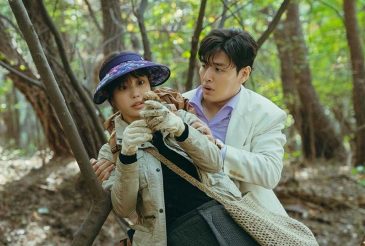 A woman and man in the woods are caught in an awkward clinch. She's fixated on a mushroom she just foraged, while he's looking at her in puzzlement