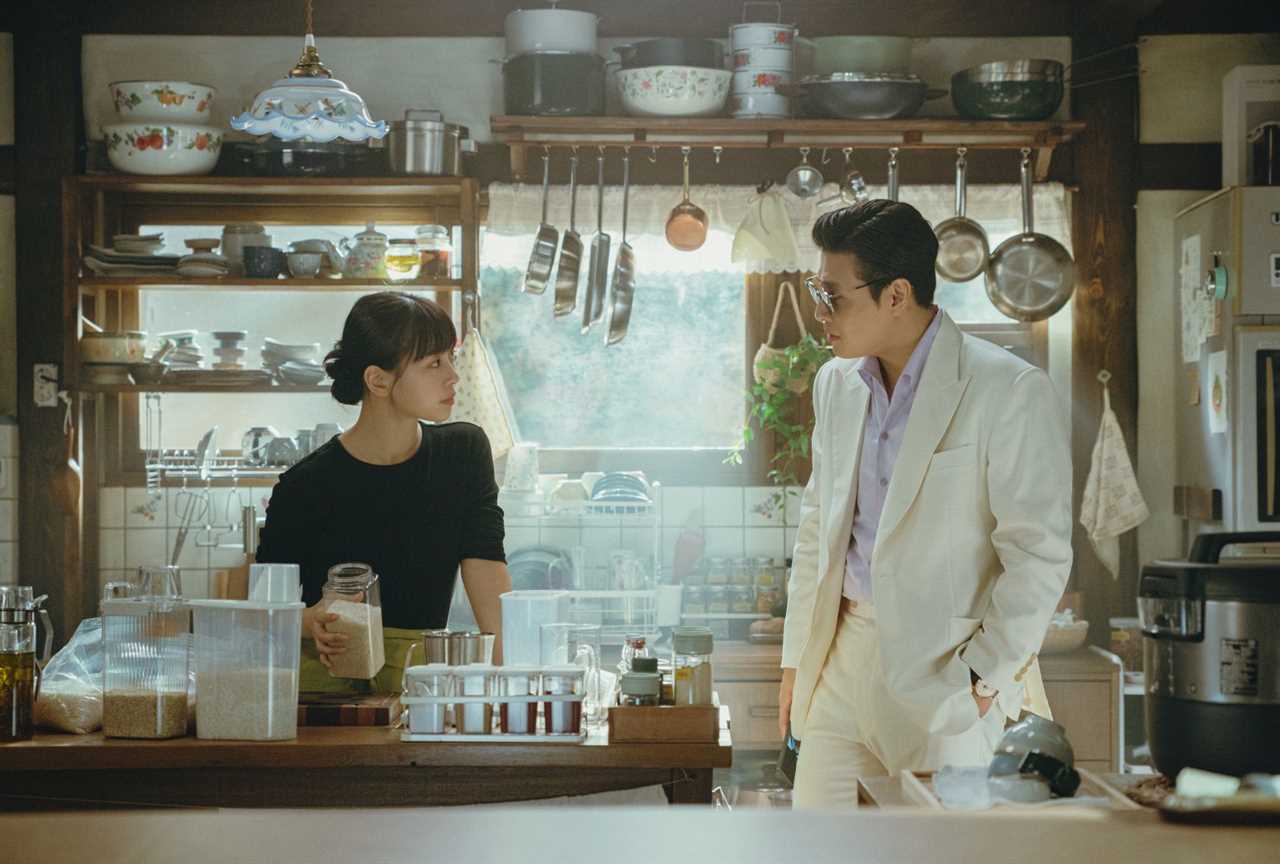 In a cosy kitchen with dishes, pots and pans stacked high on the wall, a woman in black glares at a man wearing a sharp white linen suit over a lavender shirt. He wasn't even polite enough to remove his sunglasses while speaking to her in her own home. She holds a clear container of uncooked rice grains, clearly wanting to continue cooking and not speak to him.