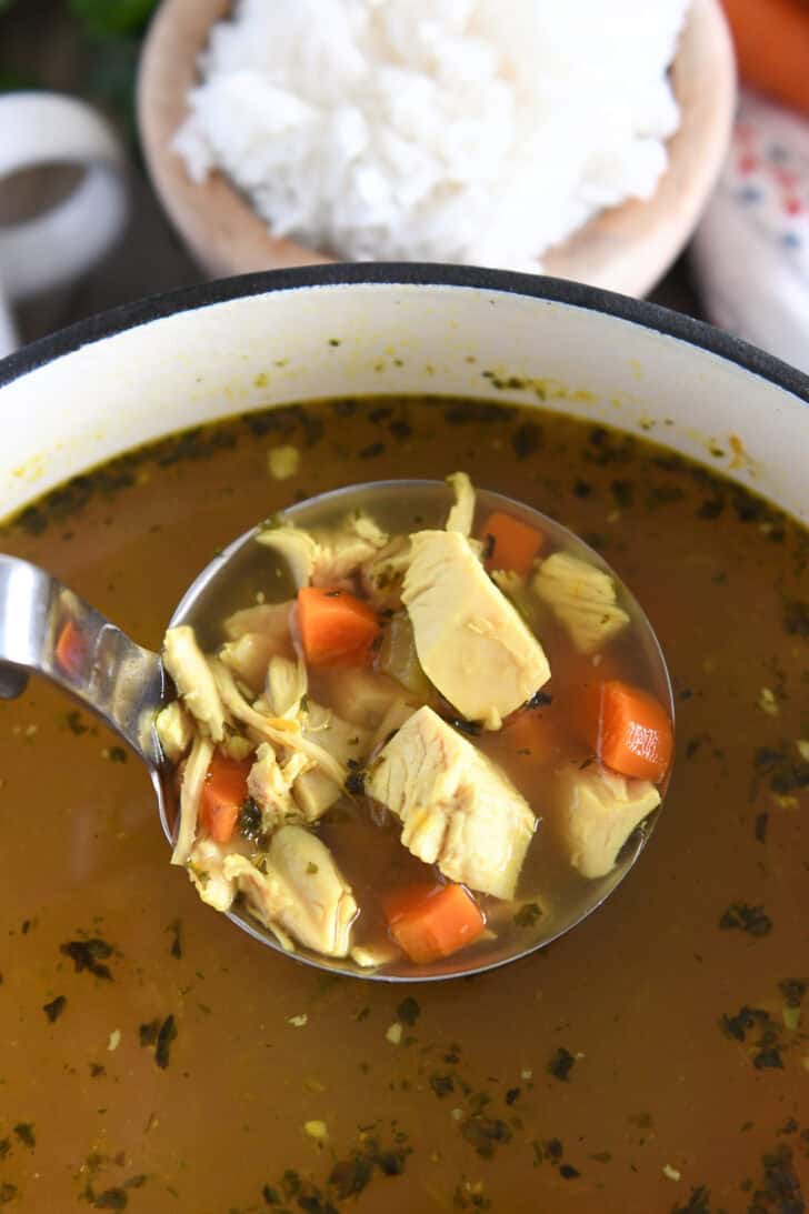Ladle lifting scoop of cooked chicken, celery and carrots out of soup pot with broth.