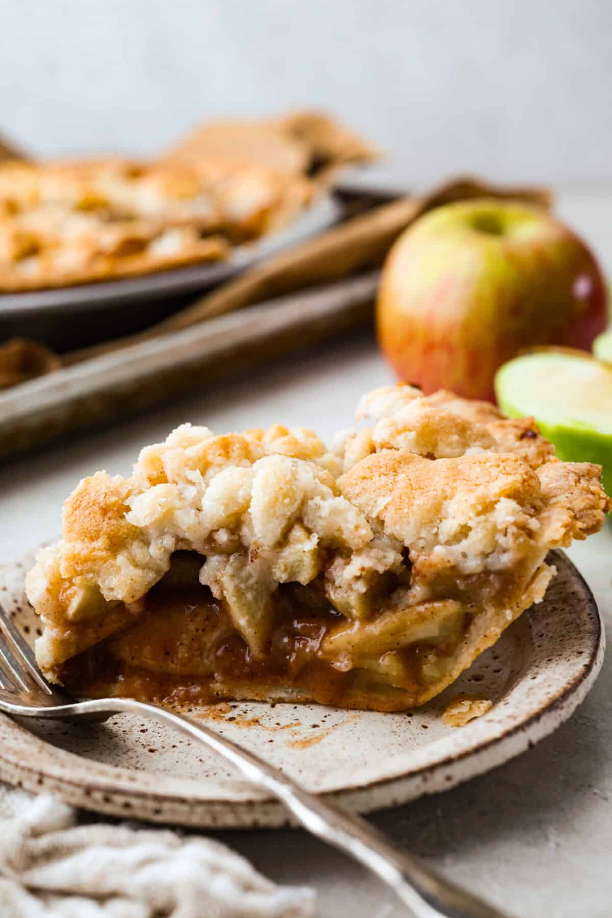 Slice of brown bag apple pie on a plate with a fork and apples in the background. 