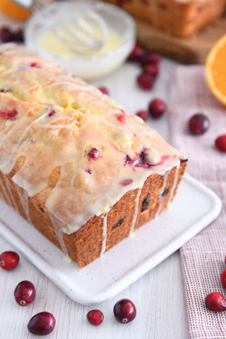 Loaf of glazed cranberry orange bread on white tray with fresh cranberries scattered around.