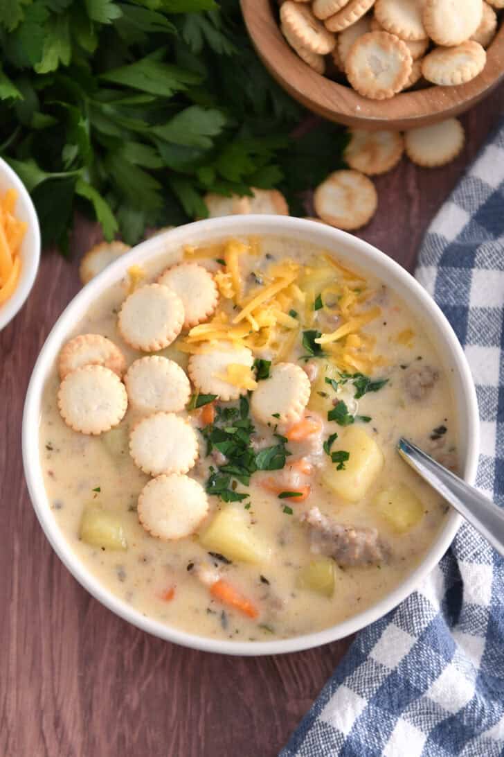 Soup bowl with sausage potato soup topped with oyster saltine crackers, fresh parsley and grated cheese and spoon on side.