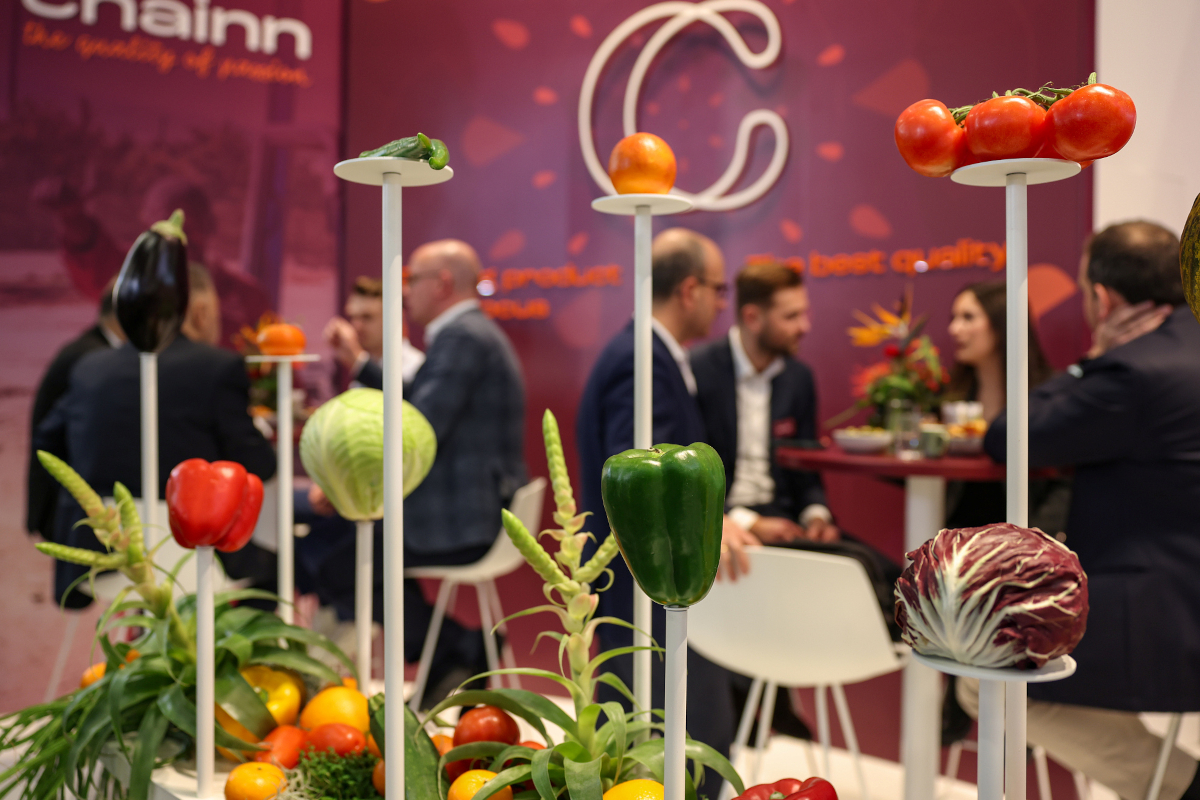 Fruit Logistica trade show display with fresh produce: tomatoes, peppers, cabbage, eggplant. People networking in background.