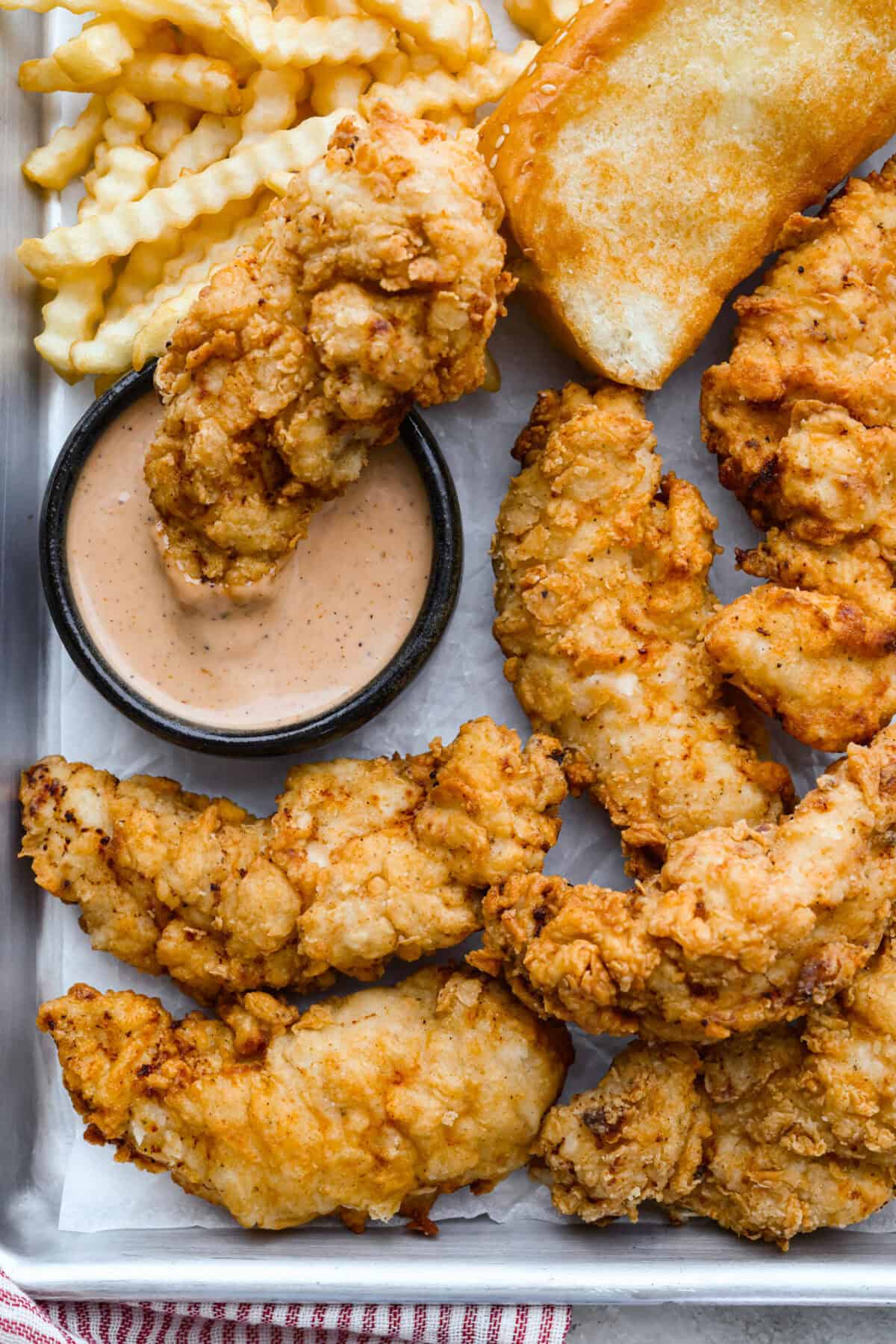 Crispy Raising Cane's chicken tenders on a plate next to crinkle cut fries, Texas toast, and a small bowl of Cane's dipping sauce. 