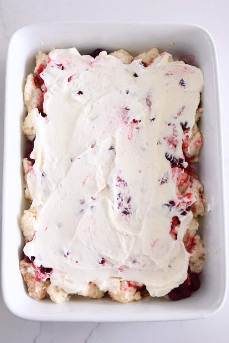 White baking dish with whipped cream layered and spread over cranberry orange layer and angel food cake cubes.