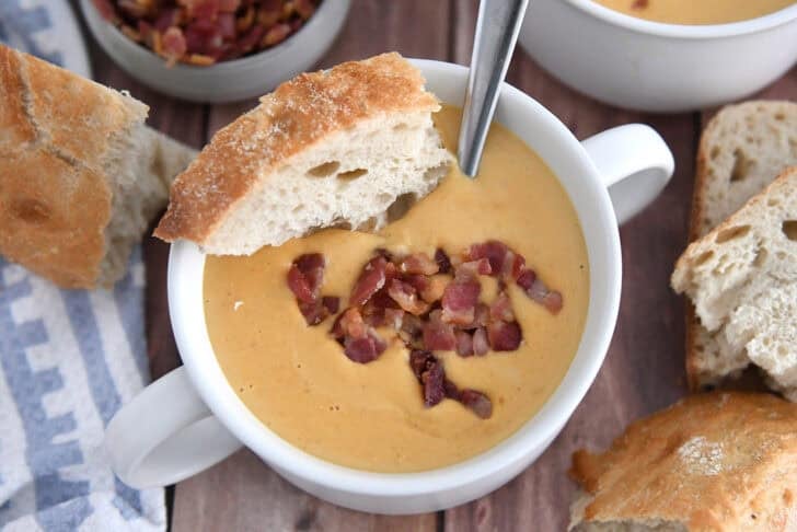 Top down view of butternut squash soup in white bowl with piece of crusty bread and cooked bacon pieces.