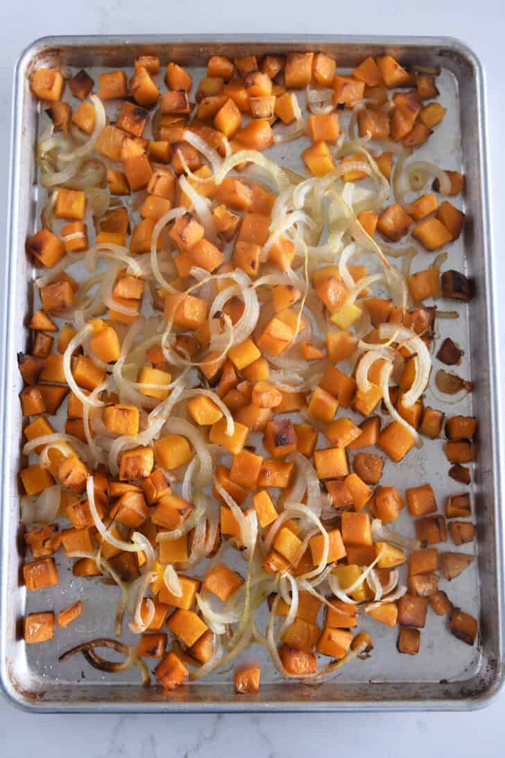 Half sheet pan with cooked butternut squash pieces and slivered onions.