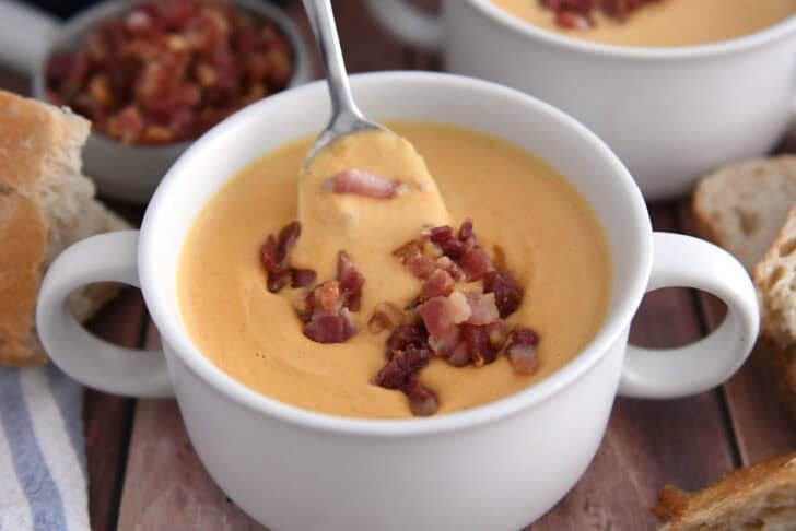 Spoon scooping bite of butternut squash soup in white bowl topped with cooked bacon pieces.