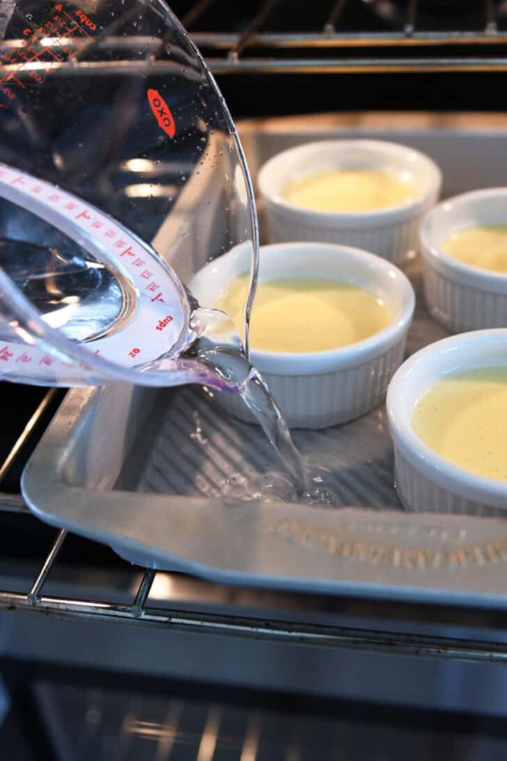 Pouring hot water into metal 9X13-inch pan in the oven with white ramekins filled with creme brulee mixture.