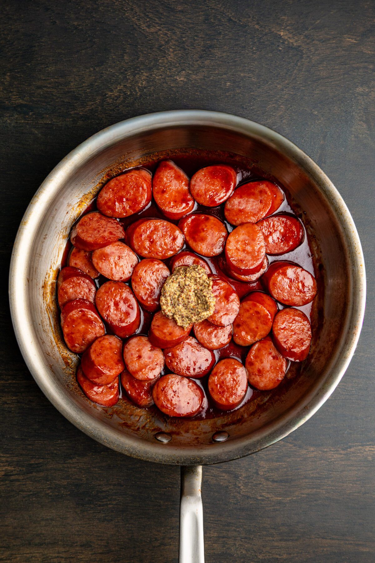 nonstick pan with mustard dolloped on top of sauce-covered kielbasa
