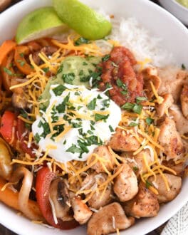 Top down view of white bowl with grilled chicken, peppers and onions, rice and limes.
