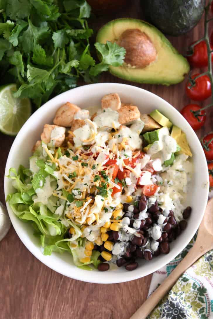 White bowl with lettuce, roasted corn, shredded cheese, tomatoes, black beans, avocados, cilantro lime rice and chicken.