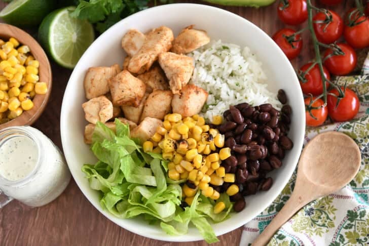 White bowl with cooked chicken, cilantro lime rice, black beans, roasted corn, and lettuce.