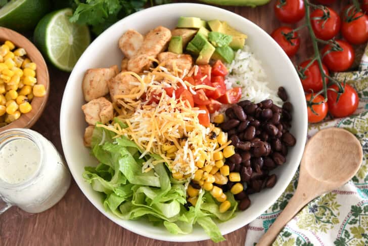 White bowl with cooked chicken, chopped avocados, tomatoes, shredded cheese, cilantro lime rice, black beans and lettuce.