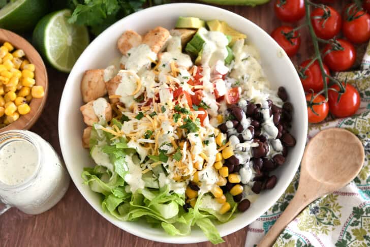 White bowl with cooked chicken, chopped tomatoes, cilantro lime rice, chopped avocados, black beans, roasted corn, and lettuce.