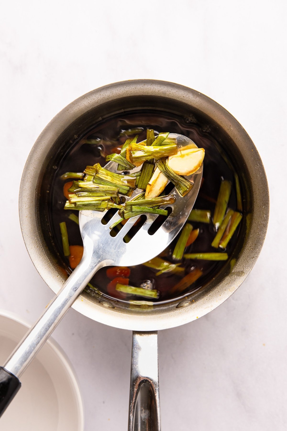 slotted spoon removing ginger, garlic, and scallions from small saucepan