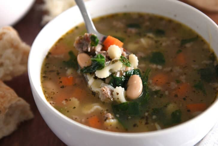 Spoon lifting bite of soup in white bowl with small pasta, spinach, carrots, white beans, and sausage.