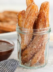 churros in glass jar with chocolate sauce and platter of churros in background.