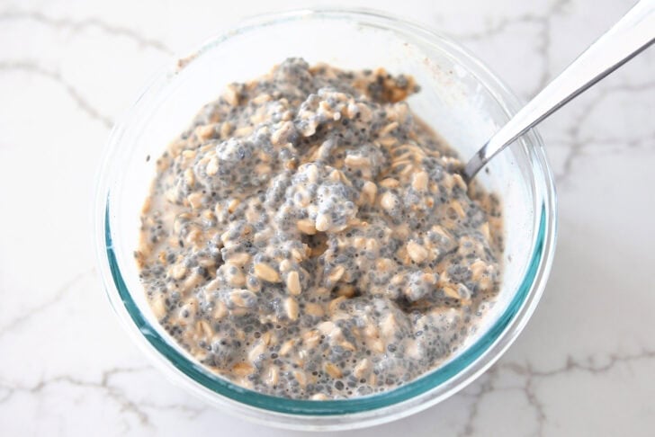 Glass bowl with spoon stirring up basil seeds, oats, and almond milk.