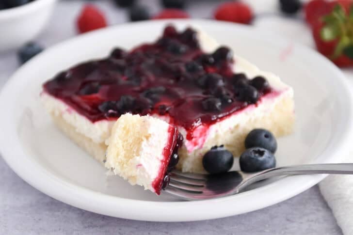 Square of white sheet cake topped with cream cheese layer and fruit topping on white plate with fresh blueberries.
