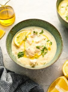 Avgolemono soup in bowl with gray striped dish towel, olive oil, and lemon wedges.