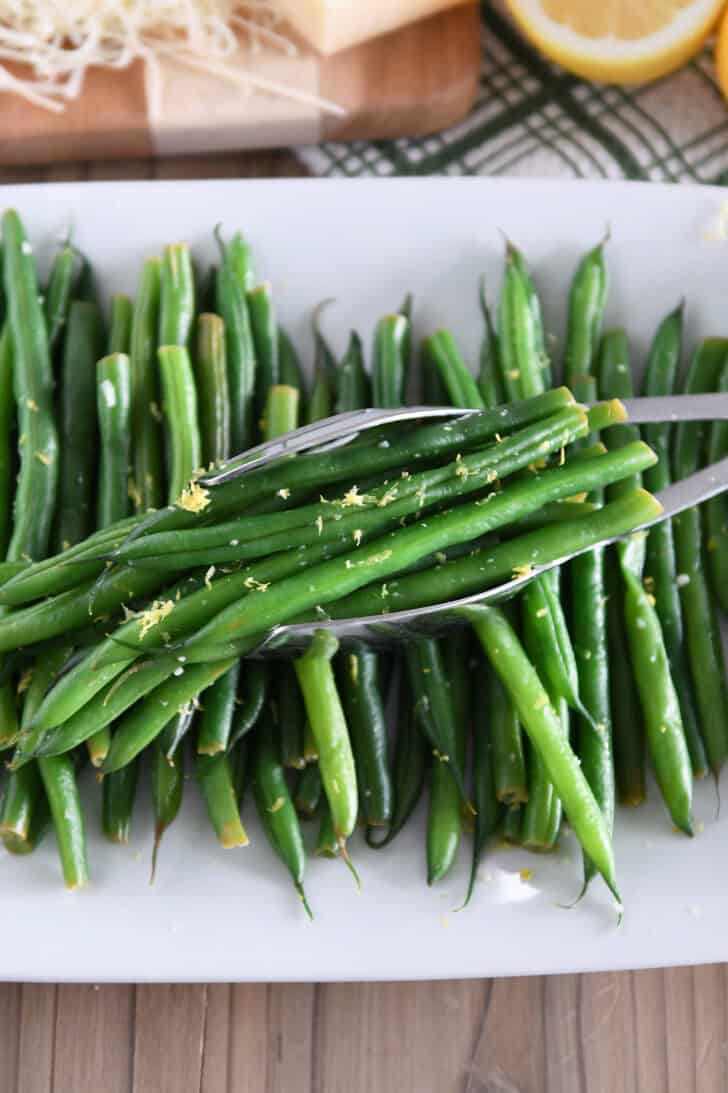 White tray with cooked green beans, lemon zest and tons scooping up portion of green beans.