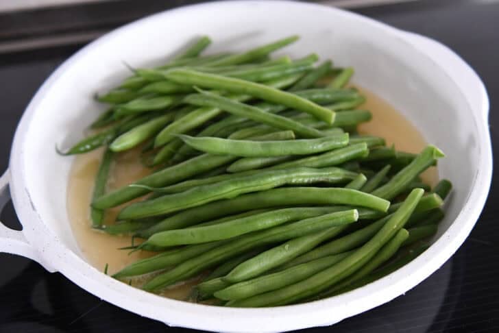 White skillet with broth and fresh uncooked green beans.