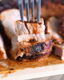 Fork piercing stack of two pieces of cooked pork tenderloin on wood cutting board.