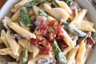 Top down view of creamy penne pasta with mushrooms, asparagus, and crumbled bacon in a white bowl.