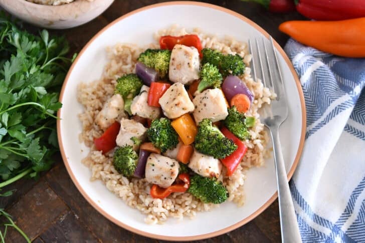 White plate with tan rim with brown rice, cooked chicken, broccoli, red onions, bell peppers, carrots and red onions.