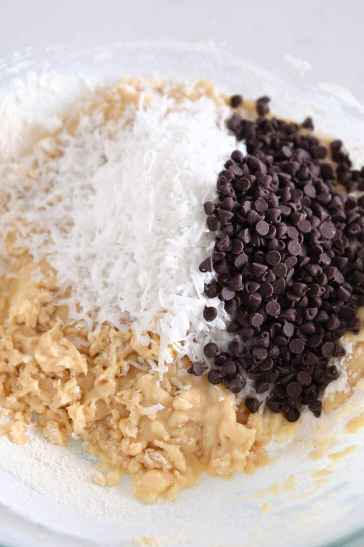 Glass bowl with muffin batter, shredded coconut and mini chocolate chips.