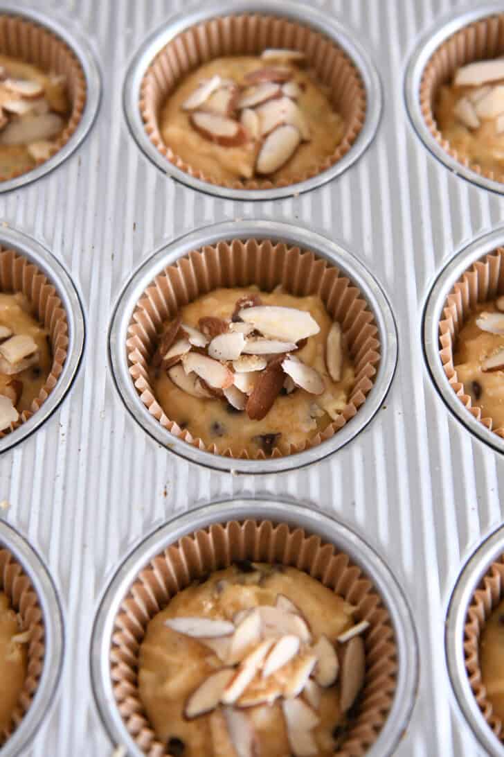 Unbaked almond coconut chocolate chip muffin batter in muffin tins topped with sliced almonds.