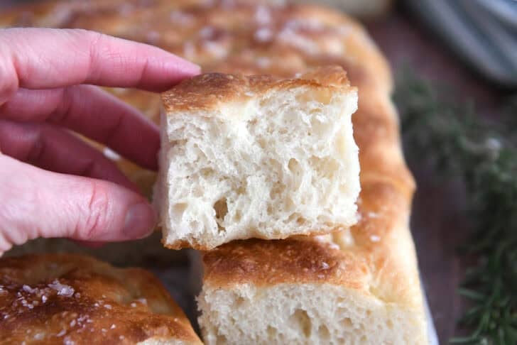 Half piece of fluffy focaccia bread on top of baked loaf of focaccia.