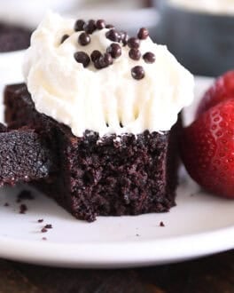 Piece of chocolate olive oil cake topped with whipped cream and crispy chocolate pearls with bite of cake on fork on white plate.