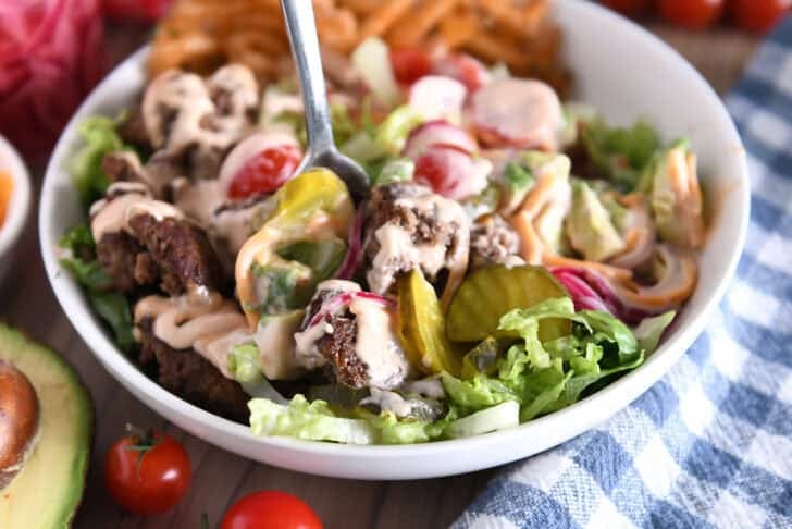 Cooked ground beef, pickles, lettuce, pickled red onions, tomatoes, fries and creamy sauce in white bowl.