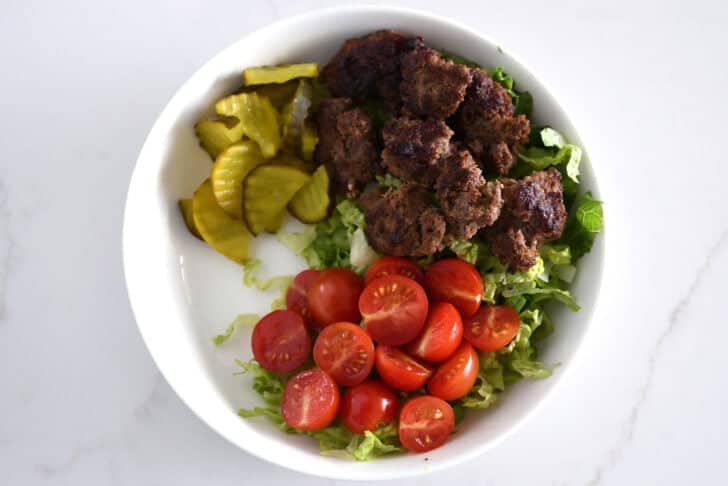 Top down view of lettuce, ground beef, tomatoes, pickles in white bowl.