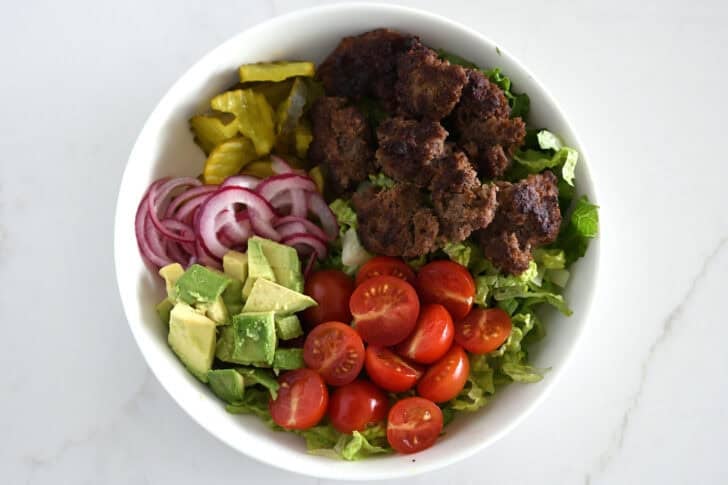 Top down view of lettuce, ground beef, tomatoes, avocados, pickled onions, pickles in white bowl.