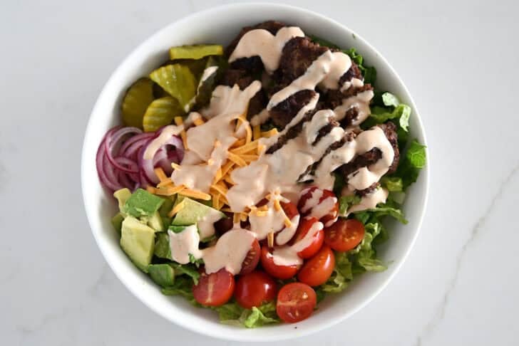 Top down view of lettuce, ground beef, tomatoes, avocados, pickled onions, pickles, shredded cheese and creamy sauce in white bowl.