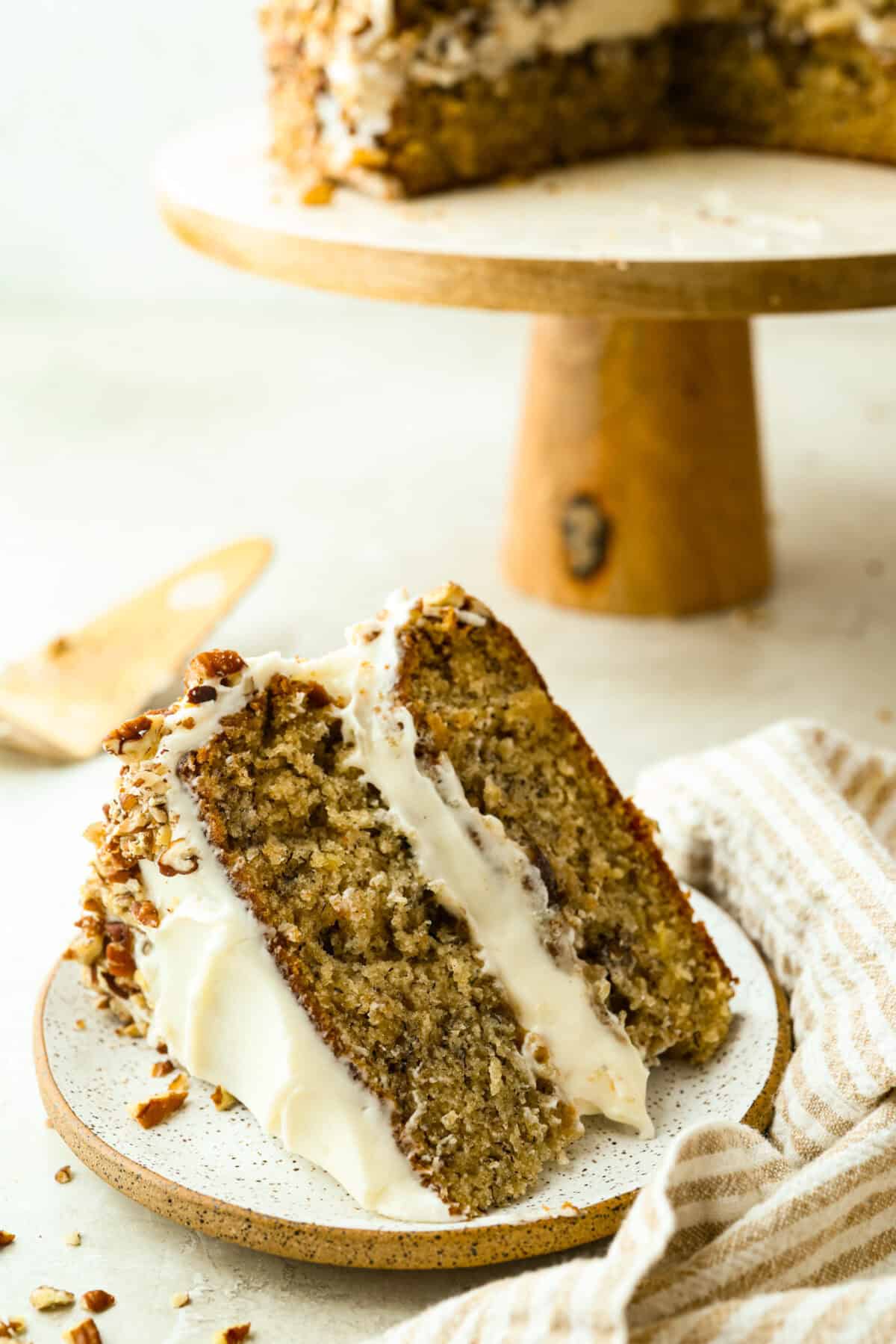 Slice of hummingbird cake on a plate next to the cake stand with the rest of the cake. 