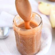 Spoon drizzling caramel sauce into glass jar with apple in the background.