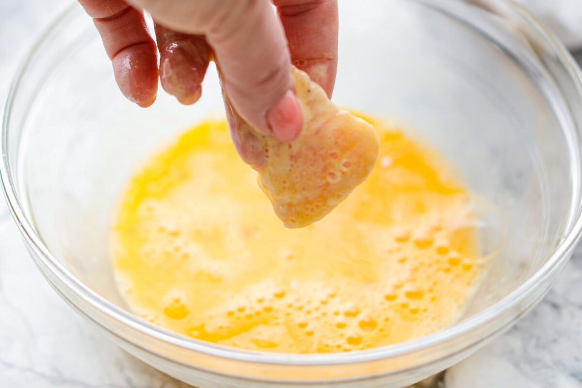 Someone dipping a piece of cornstarch coated chicken in a bowl of eggwash.