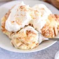 Serving of banana pudding on white plate topped with whipped cream and bite on spoon.