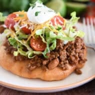 Piece of fry bread on cream plate topped with taco meat, lettuce, tomatoes, cheese, and sour cream.