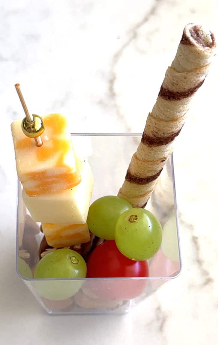 Square plastic cup with cheese skewer, blackberry, and pirouline.